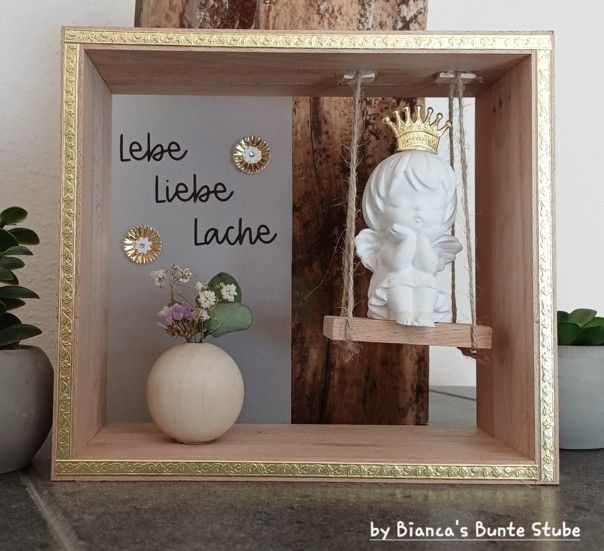 Selbst gebastelter Engel auf einer Schaukel im Holzrahmen mit Goldborte, Trockenblumen und dekorativen Blüten